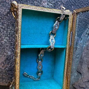 May include: An open, antique-style display box with a gold-colored frame and glass panels. Inside, a teal velvet lining and a silver and gold bracelet with blue gemstones are visible. The box has ornate detailing and a small shelf.