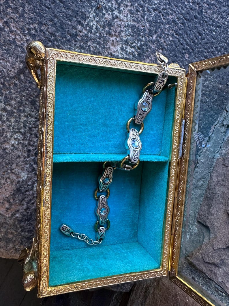 May include: An open, antique-style display box with a gold-colored frame and glass panels. Inside, a teal velvet lining and a silver and gold bracelet with blue gemstones are visible. The box has ornate detailing and a small shelf.
