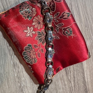 May include: A silver-toned bracelet with ornate links and blue gemstone accents rests on a red satin pouch with a floral pattern. The bracelet features detailed metalwork and a secure clasp.