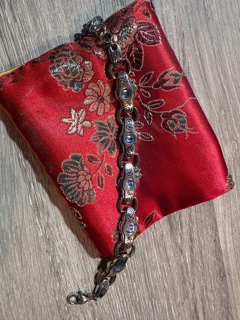 May include: A silver-toned bracelet with ornate links and blue gemstone accents rests on a red satin pouch with a floral pattern. The bracelet features detailed metalwork and a secure clasp.