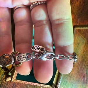 May include: Silver-toned bracelet with decorative links and a chain. The bracelet features ornate details, including floral and scroll designs. The clasp is heart-shaped. The bracelet is held by a person.