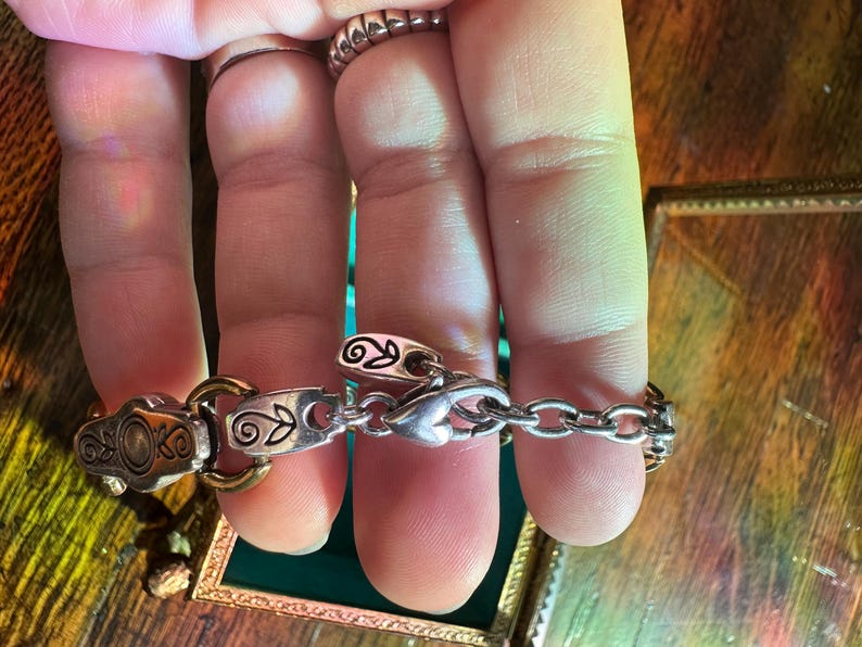 May include: Silver-toned bracelet with decorative links and a chain. The bracelet features ornate details, including floral and scroll designs. The clasp is heart-shaped. The bracelet is held by a person.