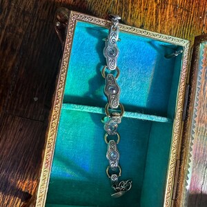 May include: A silver bracelet with oval-shaped links and decorative accents rests inside a vintage jewelry box. The box has a gold-colored frame and a teal interior. The bracelet is made of silver-colored metal and has a decorative clasp.
