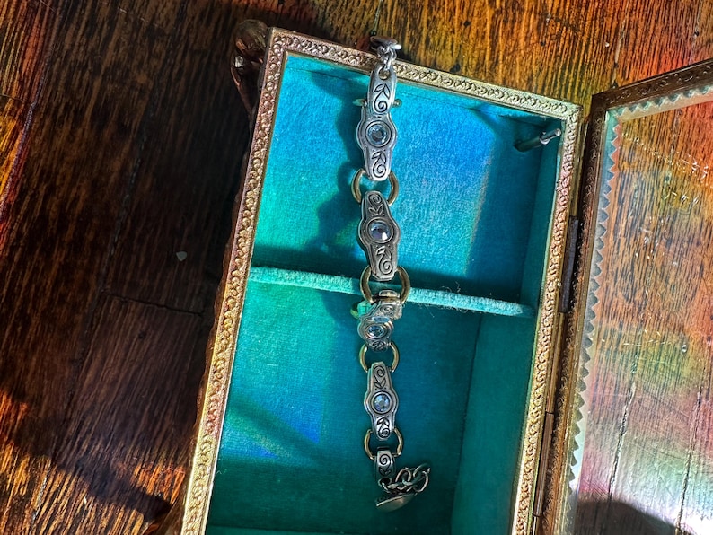 May include: A silver bracelet with oval-shaped links and decorative accents rests inside a vintage jewelry box. The box has a gold-colored frame and a teal interior. The bracelet is made of silver-colored metal and has a decorative clasp.