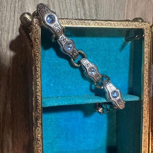 May include: A silver-toned bracelet with ornate detailing and light blue gemstones rests inside a small, antique jewelry box. The box has a gold-colored frame and a teal velvet interior, creating a striking contrast.