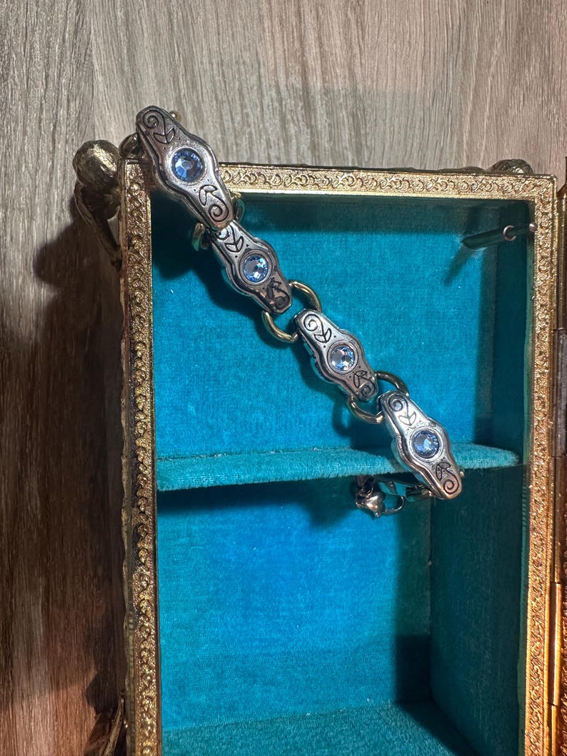 May include: A silver-toned bracelet with ornate detailing and light blue gemstones rests inside a small, antique jewelry box. The box has a gold-colored frame and a teal velvet interior, creating a striking contrast.