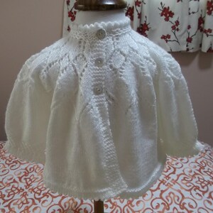 May include: A cream-colored, hand-knitted baby sweater with a round collar and three buttons. The sweater features a delicate openwork design on the yoke and sleeves. The sleeves are three-quarter length.