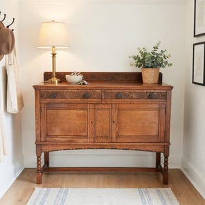 Puede incluir: Un aparador de madera de color marrón medio con dos cajones con detalles florales tallados, dos puertas de armario inferiores y patas torneadas. Una lámpara de latón con pantalla crema, un cuenco blanco y una planta verde están encima.
