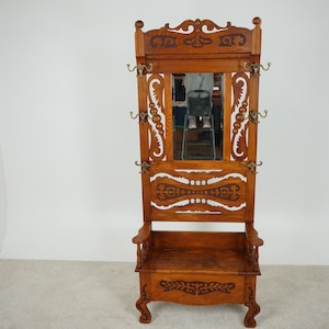 May include: A vintage wooden hall tree with a mirror, coat hooks, and a bench. The brown wood has ornate carvings and decorative accents. The hall tree is a functional piece of furniture for entryways and hallways.