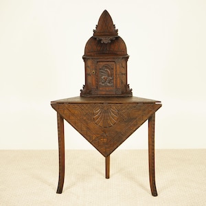 Op de afbeelding: Een donkerbruine, antieke houten kast met een driehoekige tafelvoet. De kast heeft een kleine deur met een gesneden ontwerp en een decoratieve bovenkant. De tafel heeft drie gebogen poten en gesneden details.