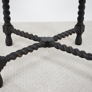 May include: Close-up of a dark brown, antique table base. The legs are intricately carved with a twisted rope design. The legs are connected by a cross-shaped support, and the feet have a rounded shape. The table base is set on a light-colored carpet.