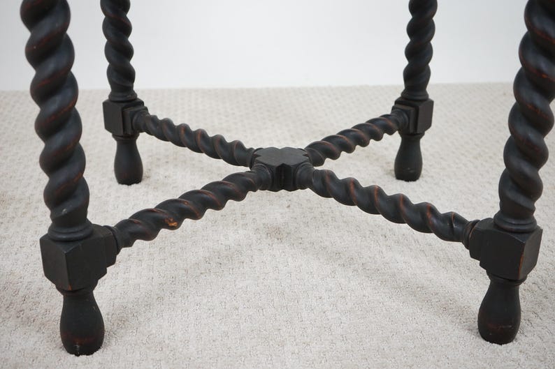 May include: Close-up of a dark brown, antique table base. The legs are intricately carved with a twisted rope design. The legs are connected by a cross-shaped support, and the feet have a rounded shape. The table base is set on a light-colored carpet.