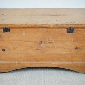 May include: A light brown wooden chest with a rectangular shape and a hinged lid. The chest has metal hardware and a curved base. The wood grain is visible, and the chest appears to be aged.