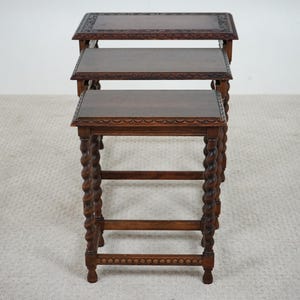 May include: A set of three nested tables made of dark brown wood. Each table features a rectangular top with a carved scalloped edge and four legs designed with a twisted barley-twist pattern, connected by horizontal stretchers at the base.