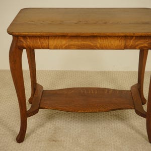 May include: A small, antique wooden table with a rectangular top and curved legs. The table has a drawer and a lower shelf. The wood has a warm, brown tone and a visible grain. The table's dimensions are approximately 30 inches long, 20 inches wide, and 28 inches high.