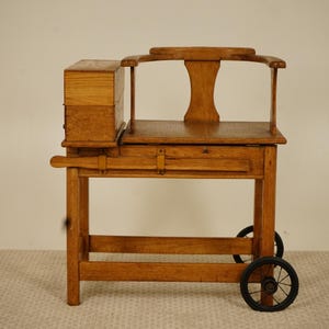 May include: A vintage wooden chair with a small box attached. The chair has a curved back and a small drawer. It is mounted on a wheeled base. The wood has a warm, natural finish. The chair's dimensions are in cm.