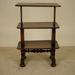 May include: A three-tiered, dark brown wooden side table. The table has a rectangular top and two shelves, supported by four carved legs. The table is approximately 76 cm tall.