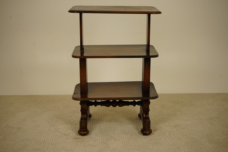 May include: A three-tiered, dark brown wooden side table. The table has a rectangular top and two shelves, supported by four carved legs. The table is approximately 76 cm tall.