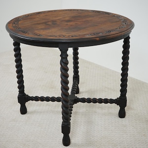 May include: A round, antique wooden side table with a dark brown finish. The tabletop features a decorative carved border and a rich, warm wood grain. The table has four dark, twisted legs connected by a lower support structure.