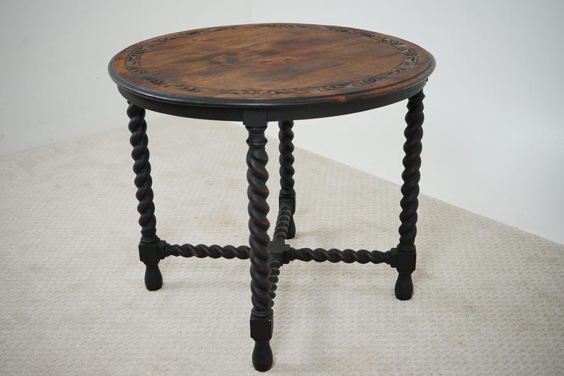 May include: A round, antique wooden side table with a dark brown finish. The tabletop features a decorative carved border and a rich, warm wood grain. The table has four dark, twisted legs connected by a lower support structure.