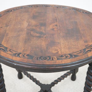 May include: A round, dark wood side table with a carved floral border and a dark finish. The table has a dark, twisted leg design. The table is approximately 24 inches in diameter and 24 inches tall.