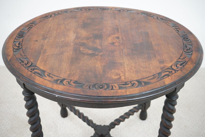 May include: A round, dark wood side table with a carved floral border and a dark finish. The table has a dark, twisted leg design. The table is approximately 24 inches in diameter and 24 inches tall.