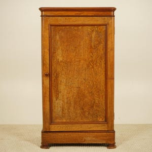 May include: A single-door wooden cabinet with a warm, medium-brown finish. The cabinet has a rectangular shape with a slightly ornate top and base. The door features a prominent wood grain pattern and a small, round knob. The cabinet is set against a neutral background.