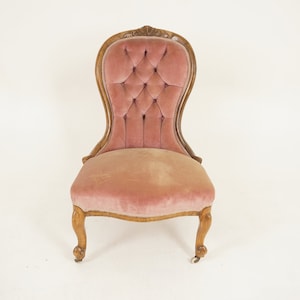Puede incluir: Una silla vintage de terciopelo rosa con respaldo capitoné y marco de madera tallada. La silla tiene cuatro patas y está sobre ruedas.