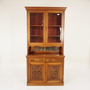 May include: A wooden cabinet with glass doors and carved details. The cabinet has two shelves and a drawer. The cabinet is in a light wood tone.
