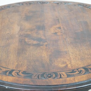 May include: A round wooden table with a dark brown finish. The tabletop features a carved border with floral and scroll designs. The table has a dark base with turned legs, showcasing a classic, antique style.