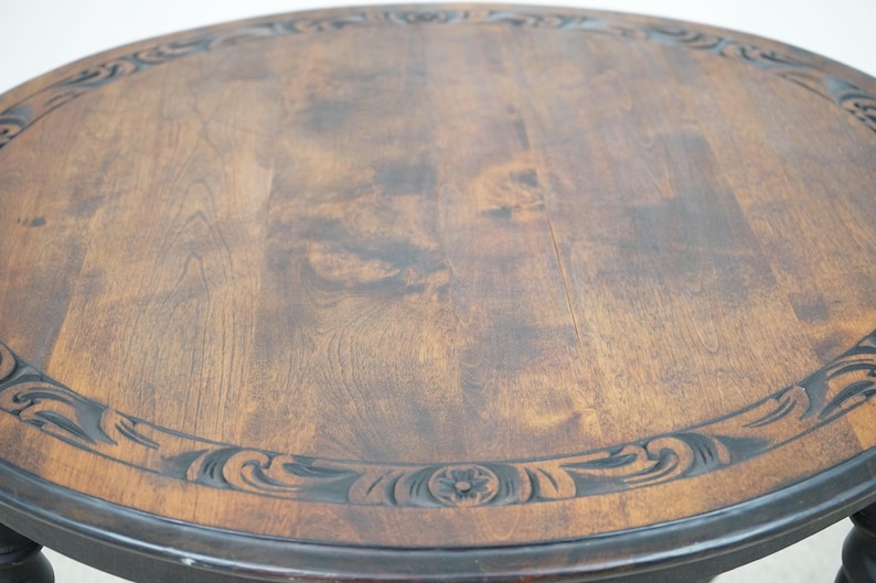 May include: A round wooden table with a dark brown finish. The tabletop features a carved border with floral and scroll designs. The table has a dark base with turned legs, showcasing a classic, antique style.
