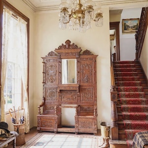 May include: A large, dark brown wooden hall tree featuring intricate carvings, a central mirror, and two integrated bench seats. The piece stands in a room with a crystal chandelier, a staircase with a red patterned runner, and tiled flooring.