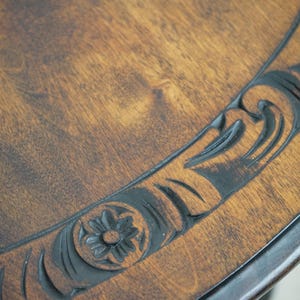 May include: Close-up of a wooden table with a dark brown finish. The table's edge features a carved floral and scroll design in a darker shade, adding a decorative element to the piece. The wood grain is visible.