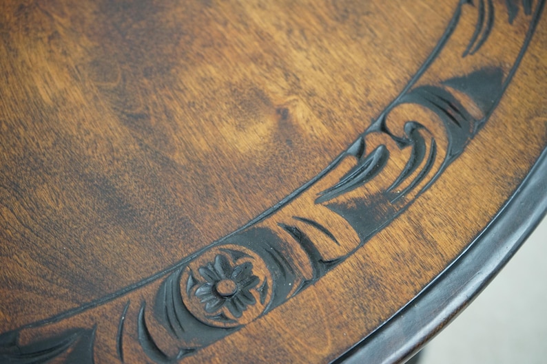 May include: Close-up of a wooden table with a dark brown finish. The table's edge features a carved floral and scroll design in a darker shade, adding a decorative element to the piece. The wood grain is visible.