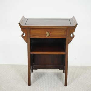 Peut inclure: Une table d'appoint en bois brun foncé avec un tiroir et deux étagères. La table a un dessus décoratif et des sculptures latérales ornées. Le tiroir comporte une poignée dorée. C'est un meuble.