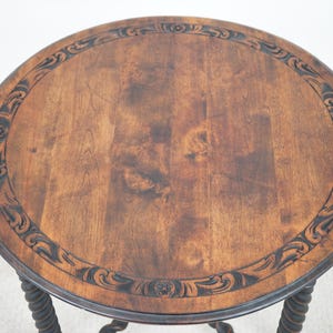 May include: A round, antique wooden side table with a dark brown finish. The tabletop features a decorative carved border and a natural wood grain pattern. The table has a twisted, rope-like leg design.