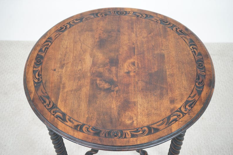 May include: A round, antique wooden side table with a dark brown finish. The tabletop features a decorative carved border and a natural wood grain pattern. The table has a twisted, rope-like leg design.