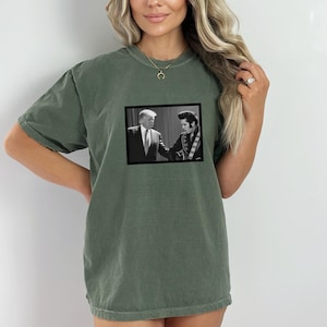 May include: A green t-shirt with a black and white photo of Donald Trump and Elvis Presley shaking hands.