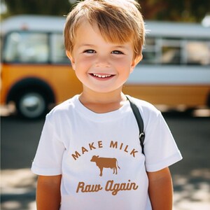 Puede incluir: Una camiseta blanca con un gr&aacute;fico marr&oacute;n que dice "Make Milk Raw Again" con una silueta de una vaca.