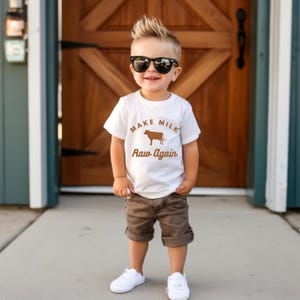 Puede incluir: Un ni&ntilde;o peque&ntilde;o con una camiseta blanca con una vaca marr&oacute;n y el texto "Make Milk Raw Again". Tambi&eacute;n lleva pantalones cortos marrones y gafas de sol negras.