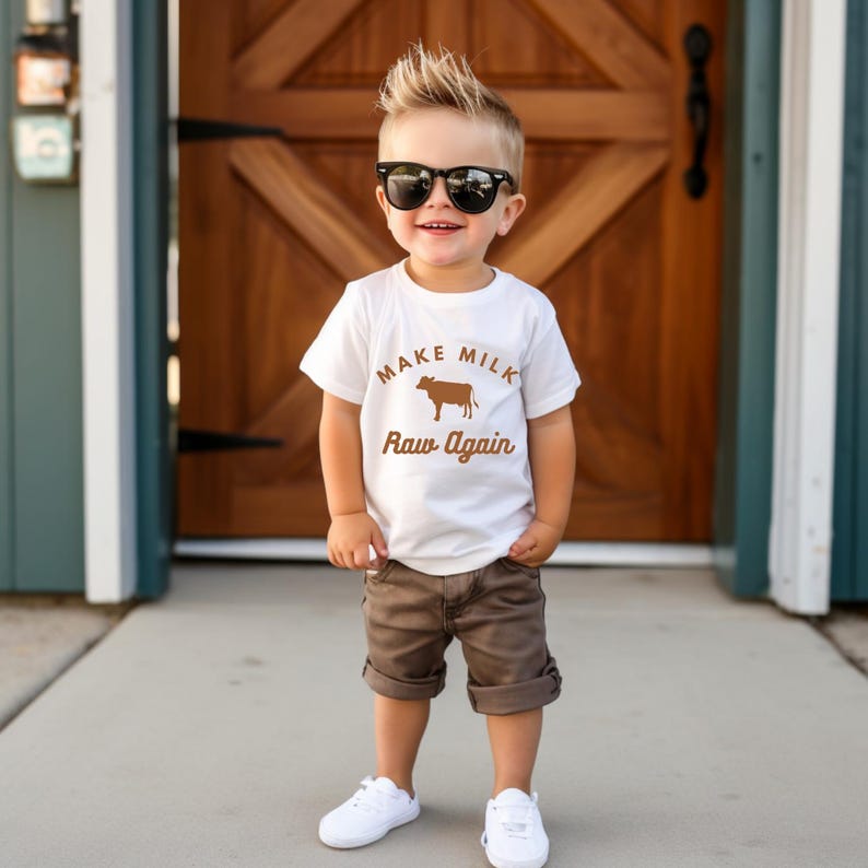 Puede incluir: Un ni&ntilde;o peque&ntilde;o con una camiseta blanca con una vaca marr&oacute;n y el texto "Make Milk Raw Again". Tambi&eacute;n lleva pantalones cortos marrones y gafas de sol negras.
