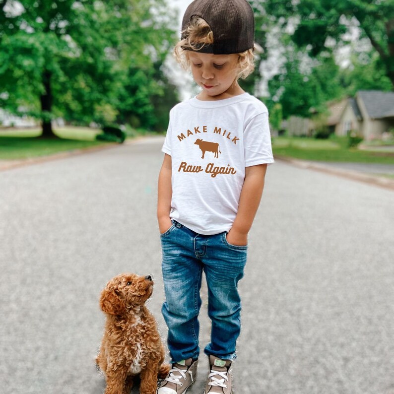 Puede incluir: Un ni&ntilde;o peque&ntilde;o con una camiseta blanca con una vaca marr&oacute;n y el texto "Make Milk Raw Again" est&aacute; de pie en un camino pavimentado con un perro marr&oacute;n a su lado.