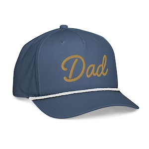 Puede incluir: Gorra de béisbol azul marino con la palabra "Dad" bordada en letras doradas. La gorra presenta un detalle de cuerda blanca a lo largo del borde y una visera curva. Un accesorio elegante para el uso diario.