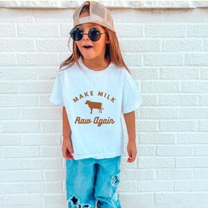 Puede incluir: Una camiseta blanca con un gr&aacute;fico de vaca marr&oacute;n y el texto "Make Milk Raw Again". Una persona joven lleva la camiseta, tiene el pelo casta&ntilde;o y lleva gafas de sol.