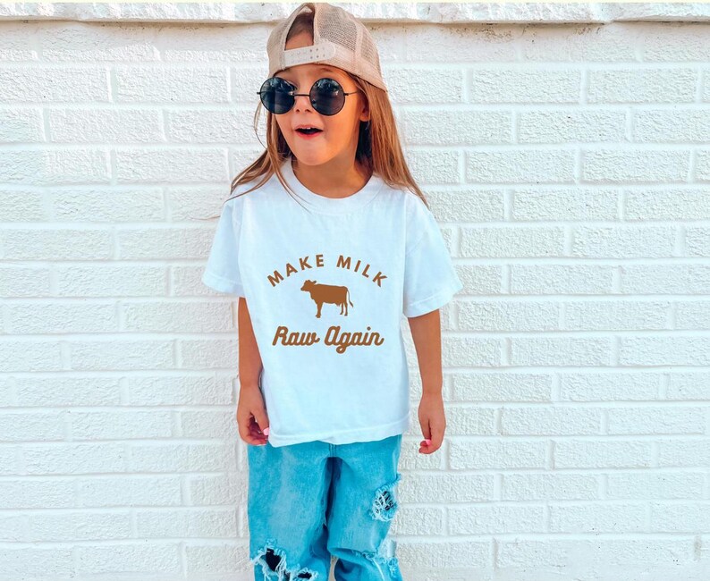 Puede incluir: Una camiseta blanca con un gr&aacute;fico de vaca marr&oacute;n y el texto "Make Milk Raw Again". Una persona joven lleva la camiseta, tiene el pelo casta&ntilde;o y lleva gafas de sol.