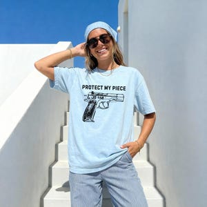 May include: Pale blue t-shirt featuring the text "PROTECT MY PIECE" and a black handgun graphic. The shirt is paired with blue and white striped trousers and a blue bandana.