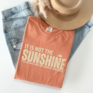 May include: A peach-colored t-shirt with the text "IT IS NOT THE SUNSHINE" in a distressed font. The shirt is paired with light-wash denim jeans and a tan fedora hat. A gold necklace is visible.