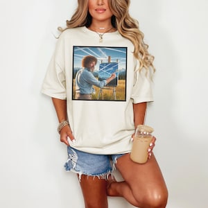 Puede incluir: Camiseta color crema con una imagen enmarcada de un pintor trabajando en un paisaje. El pintor, con afro y barba, está pintando una escena de montaña. La camiseta se combina con pantalones cortos de mezclilla.