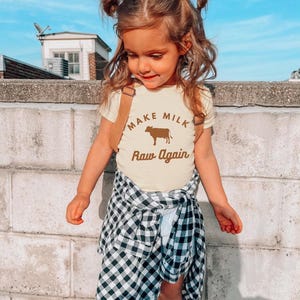 Puede incluir: Una ni&ntilde;a peque&ntilde;a con una camiseta amarilla con una vaca marr&oacute;n y el texto "Make Milk Raw Again". Lleva una camisa de cuadros negros y blancos atada a la cintura y tiene el pelo en coletas.