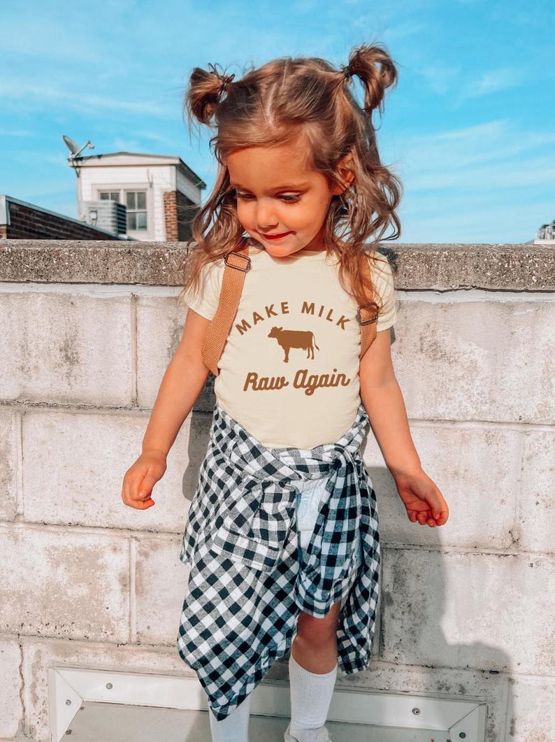 Puede incluir: Una ni&ntilde;a peque&ntilde;a con una camiseta amarilla con una vaca marr&oacute;n y el texto "Make Milk Raw Again". Lleva una camisa de cuadros negros y blancos atada a la cintura y tiene el pelo en coletas.
